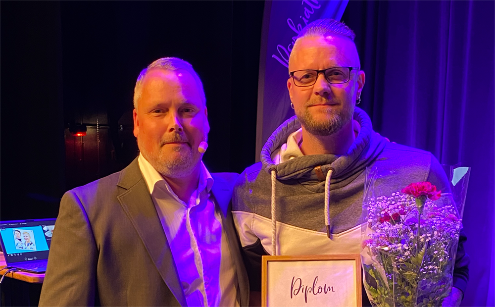Konferenciern och Martin Johansson står på en scen. Martin Johansson håller i en bukett blommor och ett diplom.