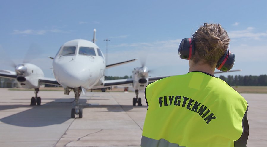 Ryggen på en person med varningsväst med texten "flygteknik" syns framför ett landande flygplan  